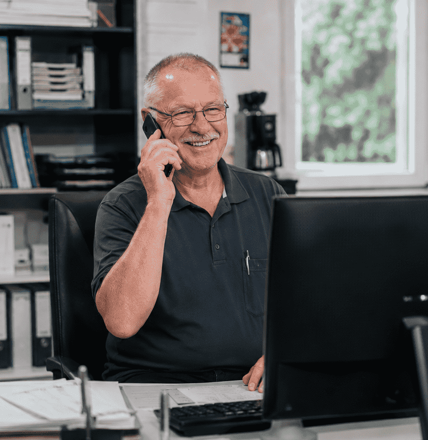 Älterer Mann lächelt am Schreibtisch beim Telefonieren vor dem Computer